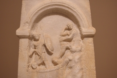 Funerary stele from Rheneia (Delos) depicting a man resting on a rock and another on a ship. The names Aulos Granios Antiokhos and Ptolemaios are inscribed on the stele, but the nature of their relationship or who is the deceased is unclear. The meaning of the scene is also unclear, but suggests that at least one of the men died at sea. Dated to around 100 BCE. Szépművészeti Múzeum.